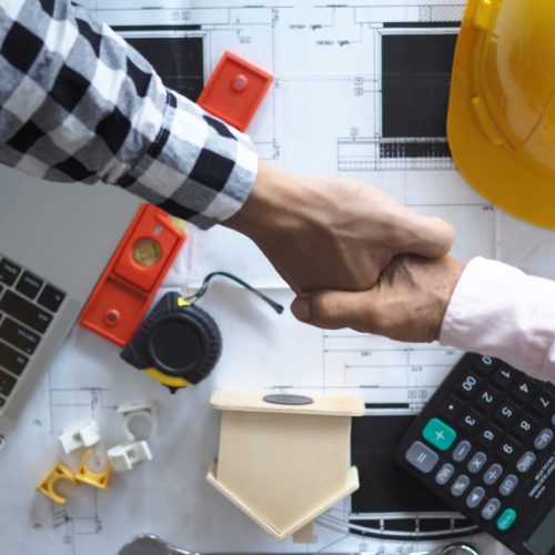Top view of a handshake above a calculator, floor plans, and construction tools.